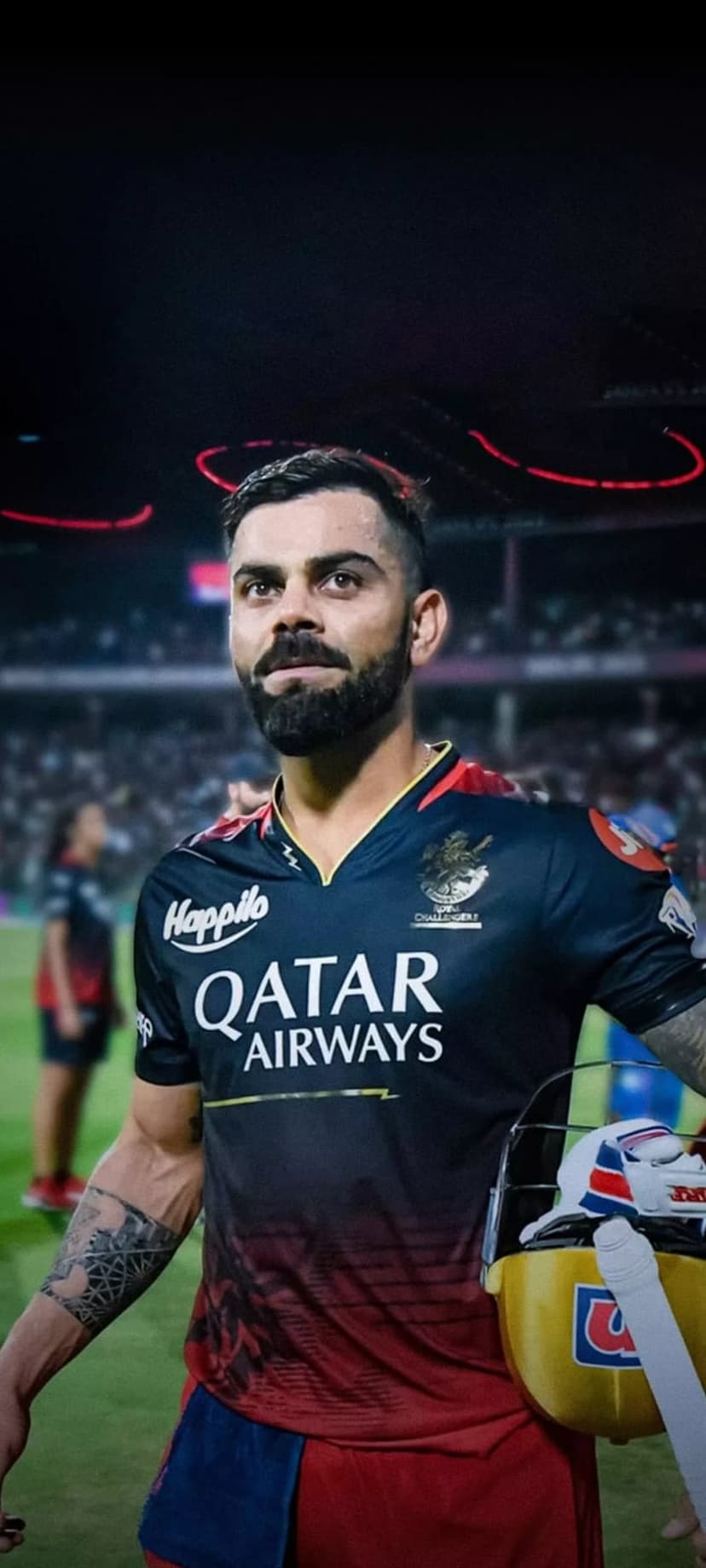 Virat Kohli in a red RCB jersey holds a cricket helmet before a blurred stadium backdrop with glowing lights.