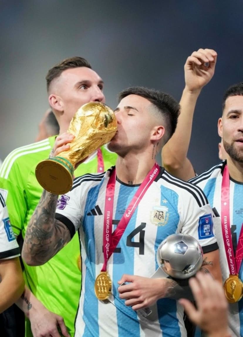 Enzo Fernández kisses the golden World Cup trophy alongside cheering Argentina teammates wearing gold medals.