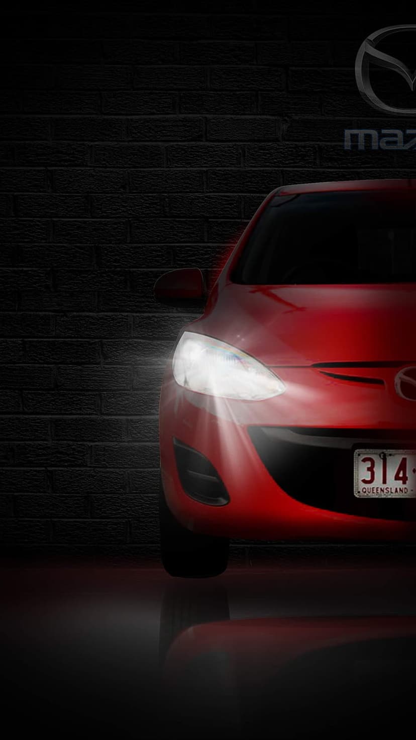 Red Mazda sports car with glowing headlights parked against a dark brick wall with a glossy ground reflection.