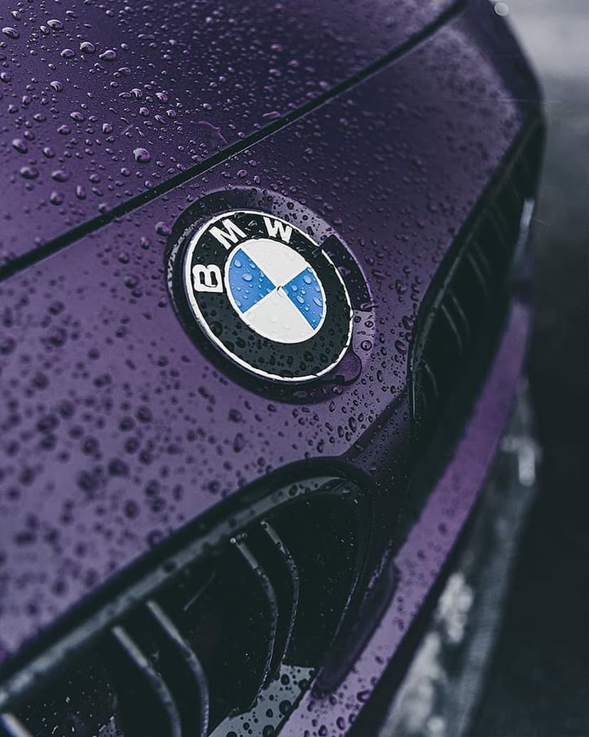 Silver BMW logo and grille textured with rain droplets on a deep violet automotive surface in close-up.