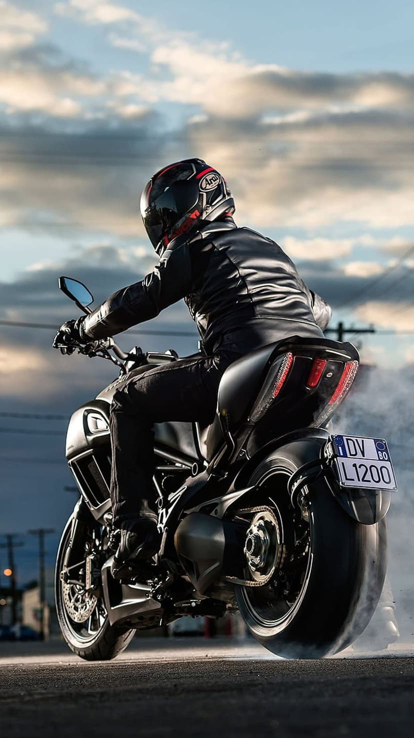 Ducati Diavel rider performs a rear wheel burnout with thick white smoke against a dramatic urban sky.