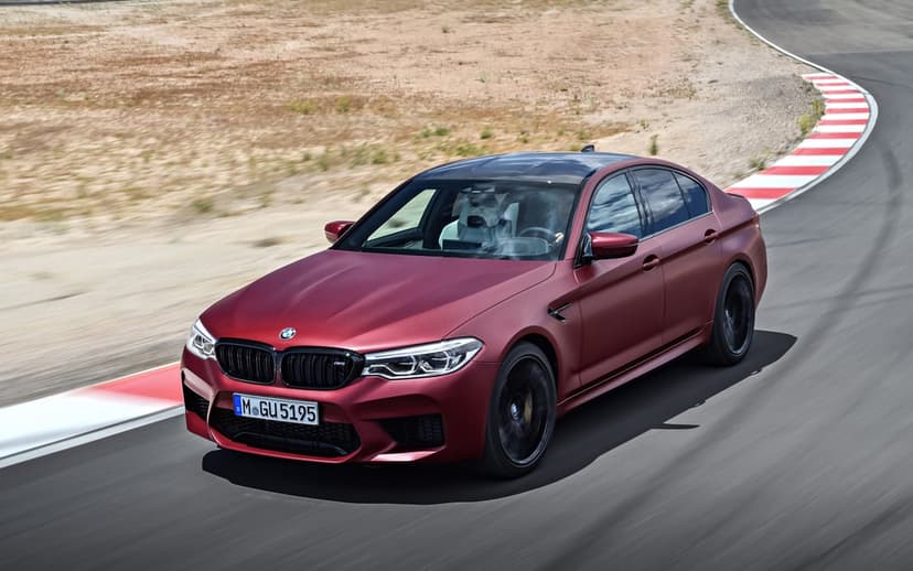 Matte red BMW M5 sports sedan driving through a curve on a racetrack with red and white kerbs in bright daylight.