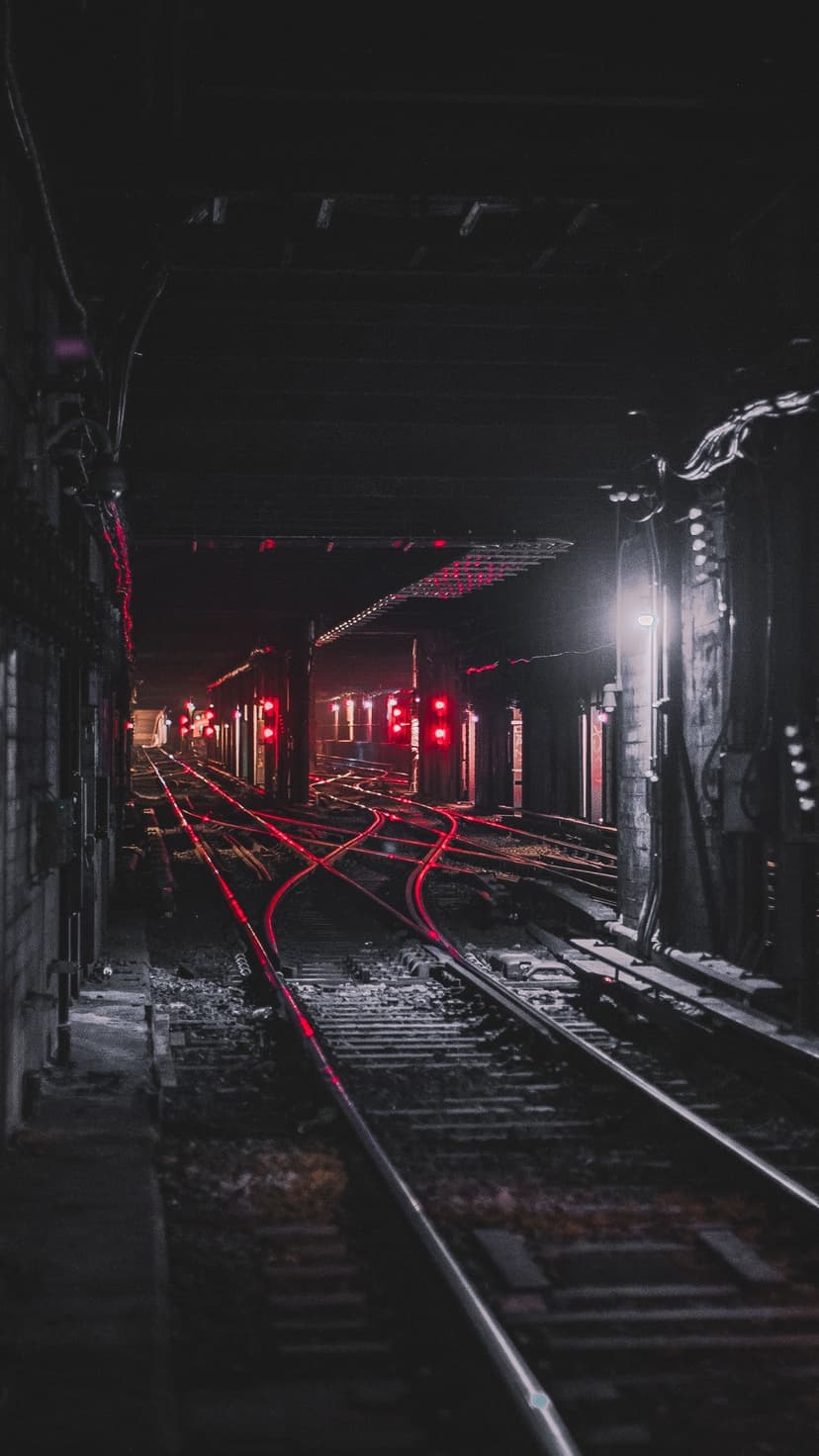 Subway tracks recede into a dark tunnel under glowing red signal lights reflecting on damp, industrial floors.