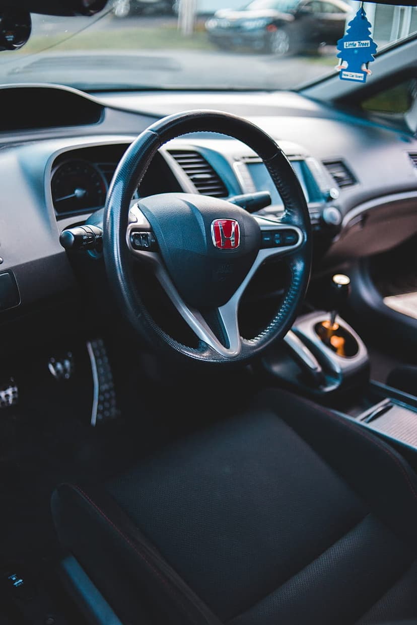 Honda steering wheel with a red logo and blue air freshener hangs in a dark car interior with a gear shifter.