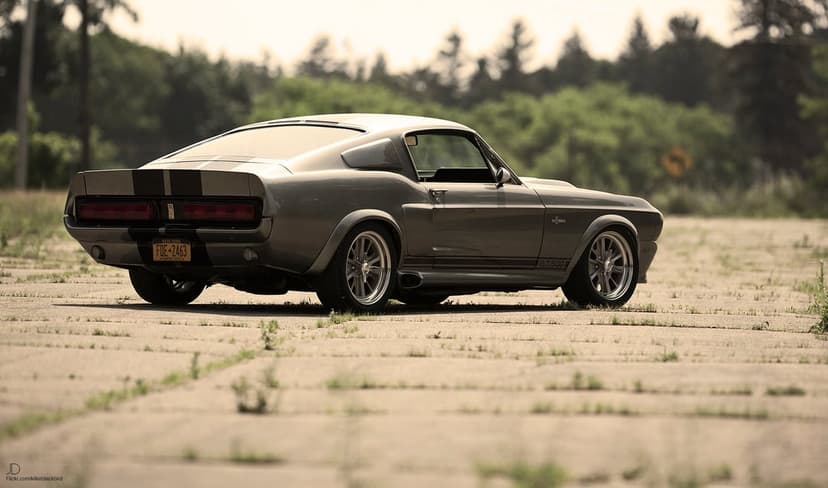 Rear view of a gray Shelby GT500 Eleanor with black stripes on a weathered airfield with blurred trees.