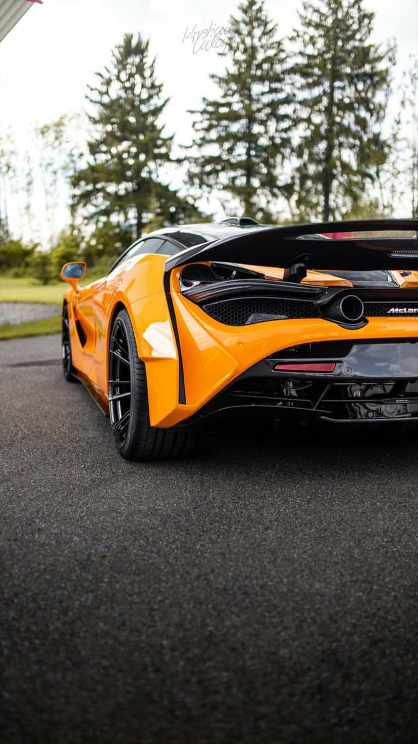 Orange McLaren 720S supercar rear view with visible exhaust and spoiler on a road under an overcast sky.