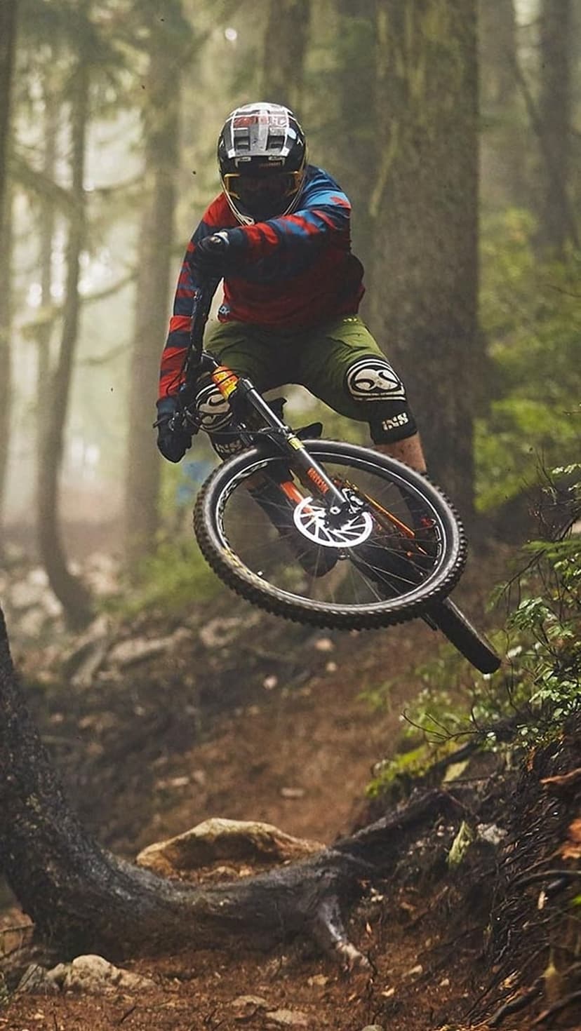Mountain biker wearing a full-face helmet jumps mid-air over a log on a foggy, moss-covered forest trail.
