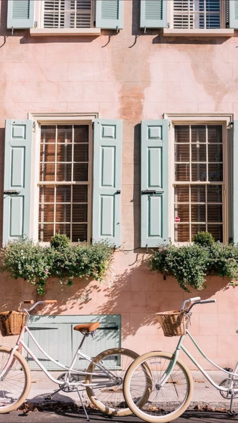 Pastel pink building with light blue shutters and two vintage bicycles parked in front of a green door in sunlight.