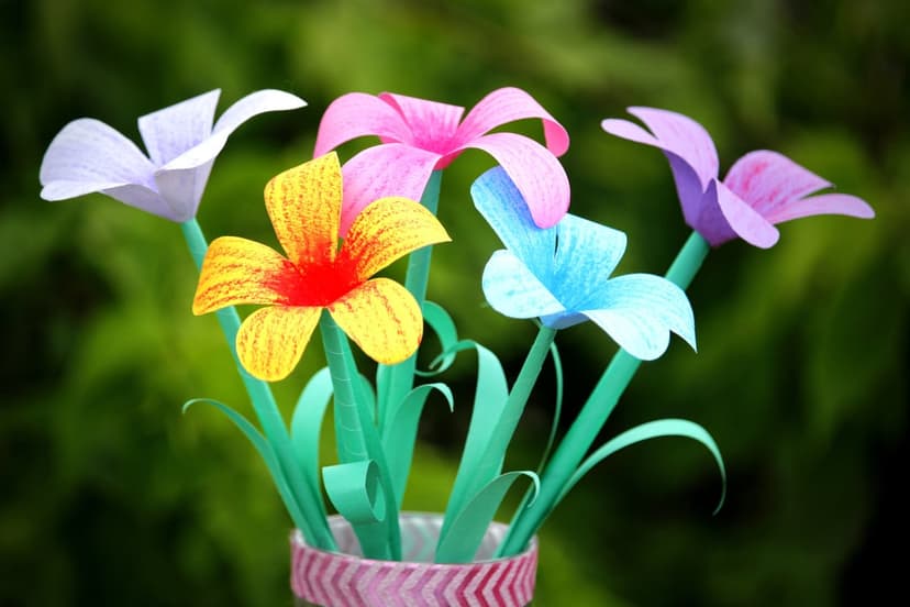 Vibrant paper flowers in yellow, pink, and blue stand in a patterned vase before a soft green bokeh background.