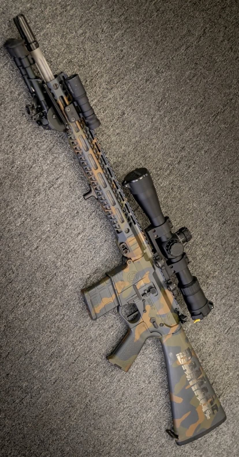AR-15 rifle with brown and tan camouflage, magnified scope, and bipod resting on a flat surface in portrait view.