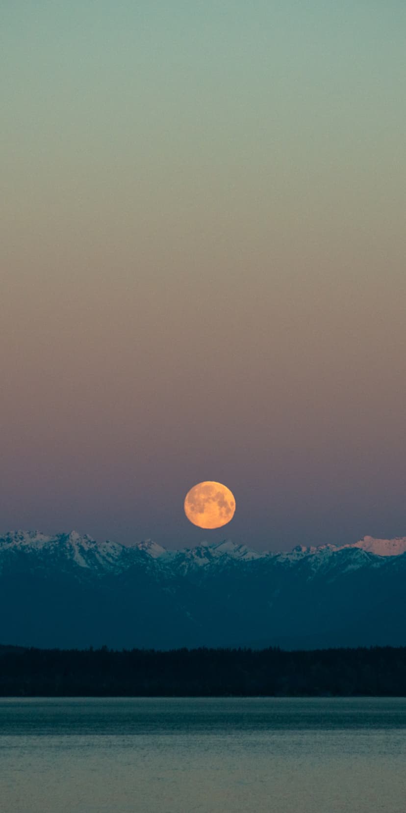 Large orange moon hovering over snowy mountain peaks and a still lake with a purple and blue gradient sky.