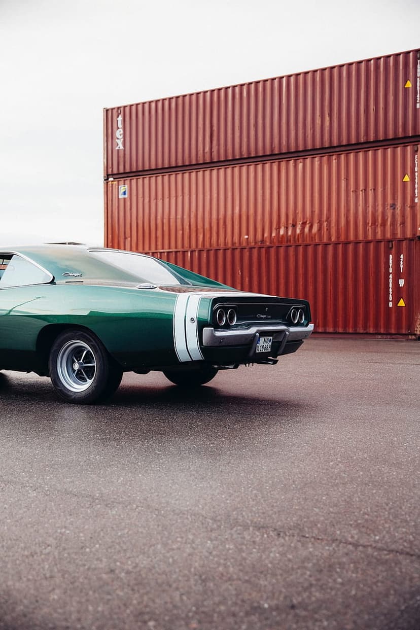 Green vintage muscle car with white stripes parked on wet asphalt near red shipping containers in low angle.