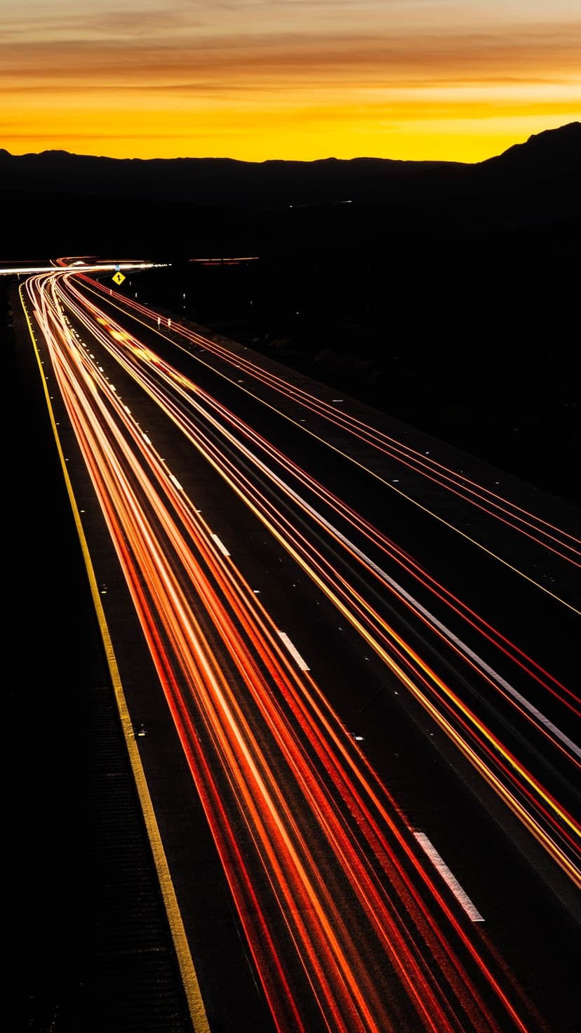 Red and white vehicle light trails curve through a dark desert landscape under a fading sunset horizon.