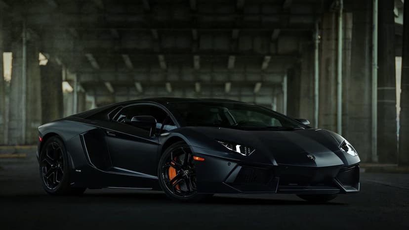Matte black Lamborghini Aventador with orange calipers under a concrete bridge with glowing white headlights.