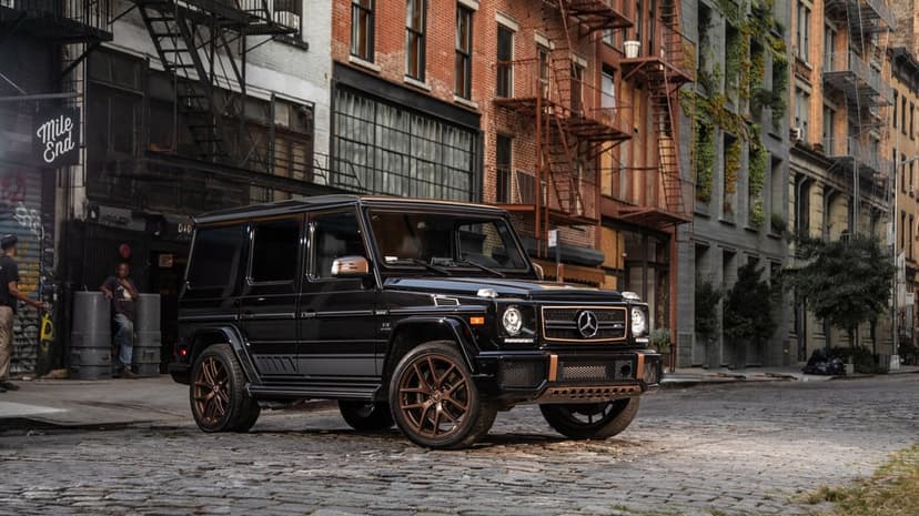 Black Mercedes G-Class SUV with bronze wheels parked on a cobblestone street in front of brick city buildings.