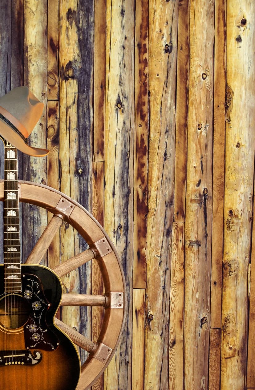 Acoustic guitar rests against a weathered barn wall beside an antique wooden wagon wheel in a rustic setting.