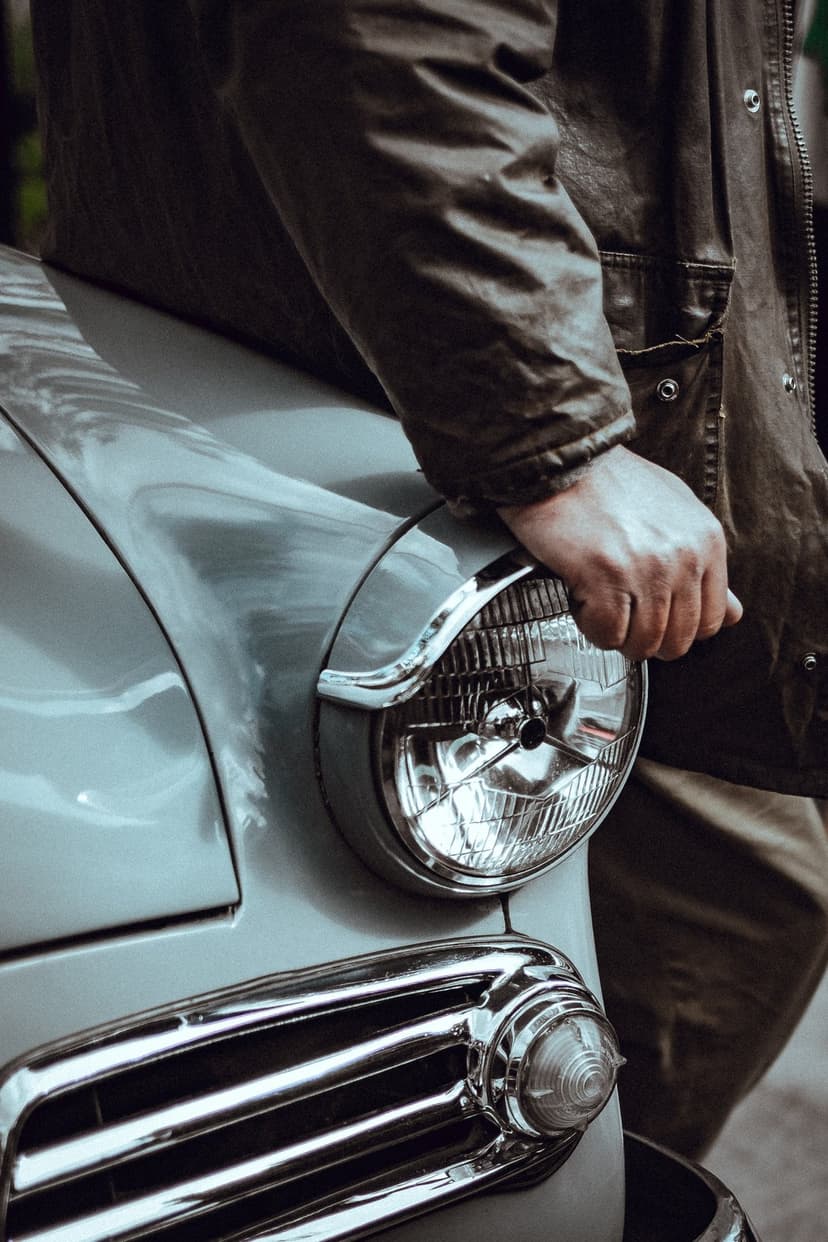 Man's hand in a dark jacket rests on the circular chrome headlight of a classic light blue vintage automobile.