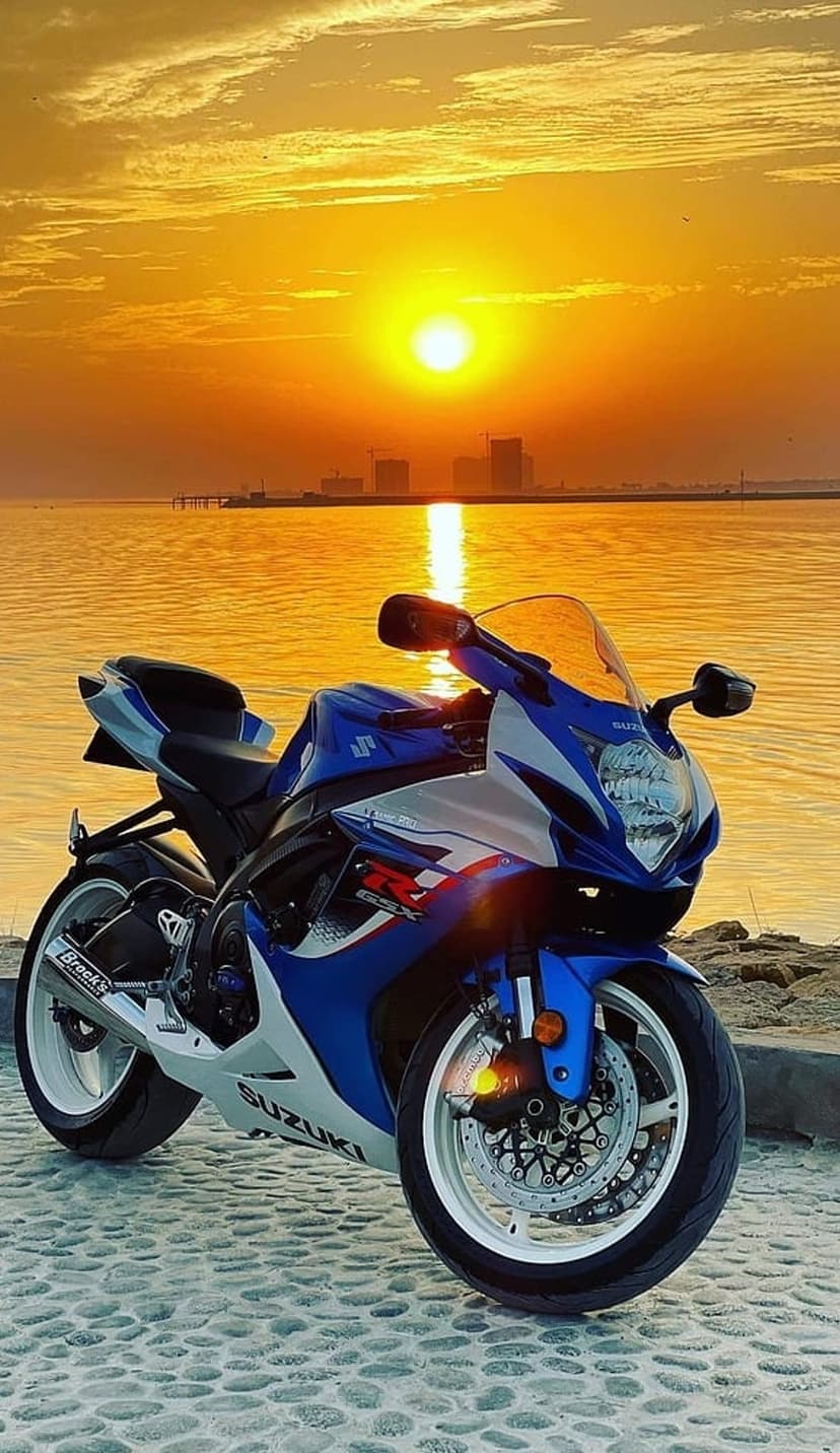 Suzuki sportbike in blue and white on a cobblestone beach during a golden sunset with a distant city skyline.