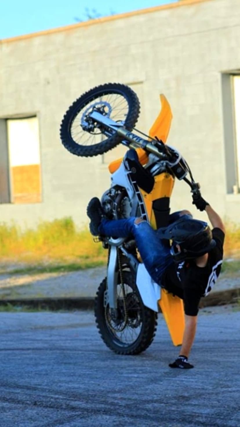 Motorcyclist in a helmet executes a steep wheelie on a dirt bike against a blurred field and building.