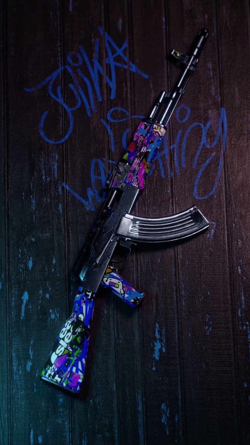 AK-47 rifle with colorful pixel art skin lying on dark wooden planks with scattered blue graffiti paint splatters.