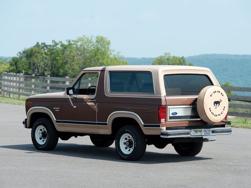 Vintage brown and tan Ford Bronco XLT parked on a road with a spare tire cover and mountain backdrop.