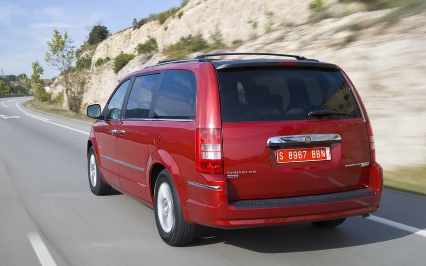 Rear view of a red Chrysler Grand Voyager minivan driving fast on a sunlit highway past rocky hills.