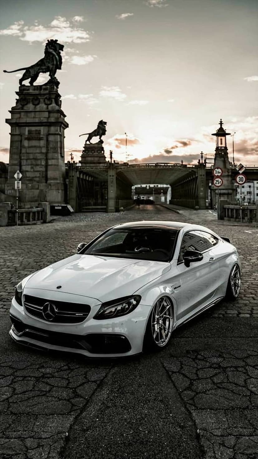 White Mercedes-AMG C63 Coupe parked on a cobblestone street near stone lion statues during a soft golden sunset.