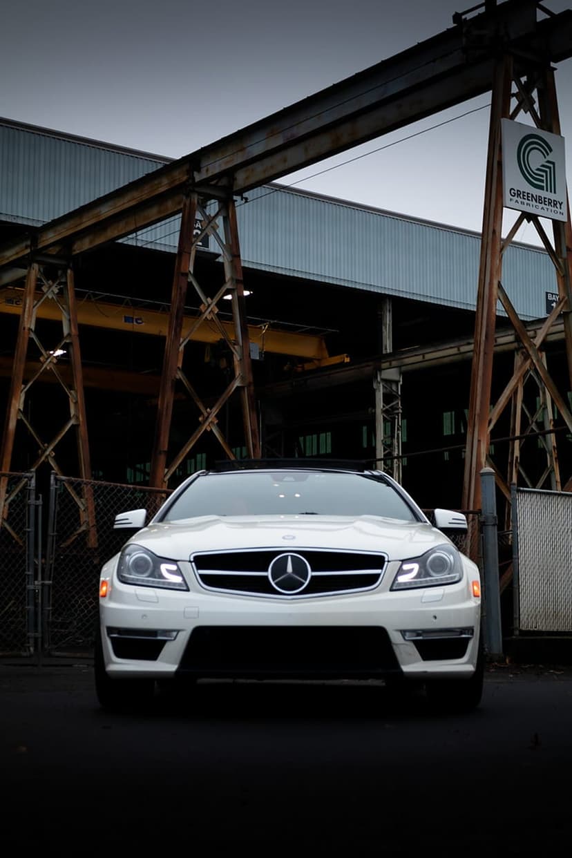 White Mercedes-Benz C-Class coupe with glowing headlights parked in front of a dark, rusted industrial factory wall.