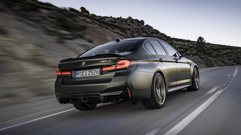 Rear view of a matte green BMW M5 CS driving fast on a mountain road with glowing red taillights.