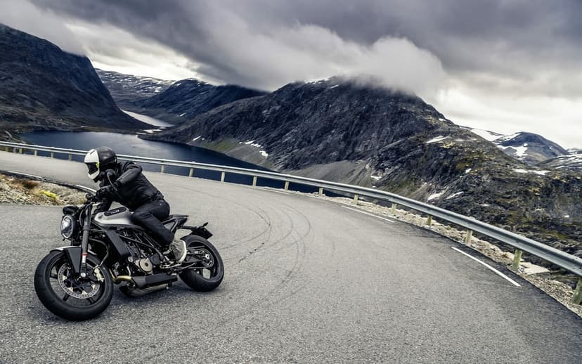Motorcyclist in black leather leans a silver bike on a winding road overlooking mountains and a blue lake.