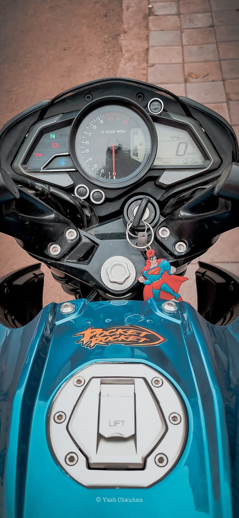 Superman keychain hangs from a teal motorcycle ignition beside a digital speedometer and analog tachometer.