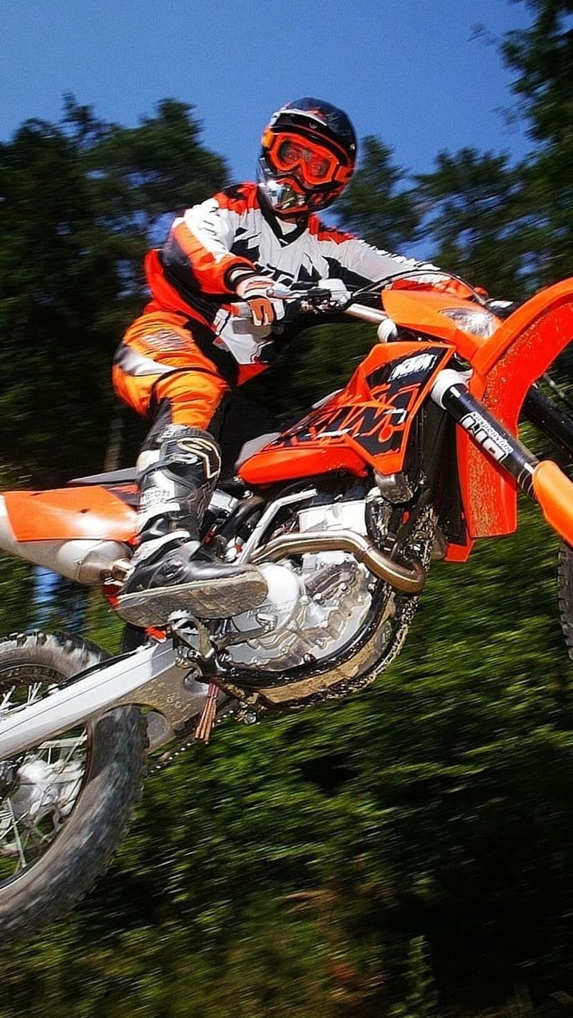 Motocross rider in orange gear jumps a dirt bike against a blue sky and blurred green trees in mid-air flight.