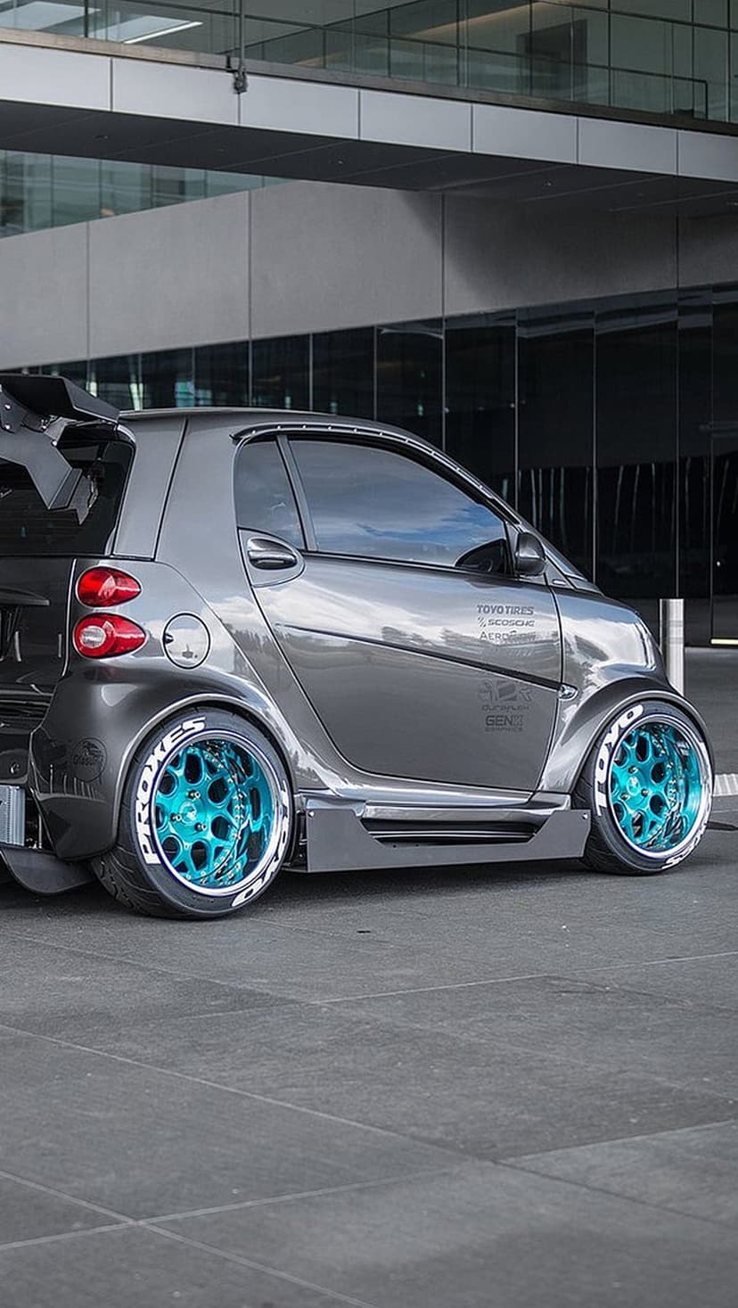 Gray customized microcar with bright teal wheels and a rear spoiler parked on tiles near a glass building.