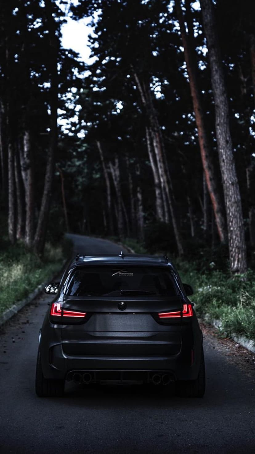 Matte black SUV with glowing red taillights driving through a dark, dense forest on a winding asphalt road.