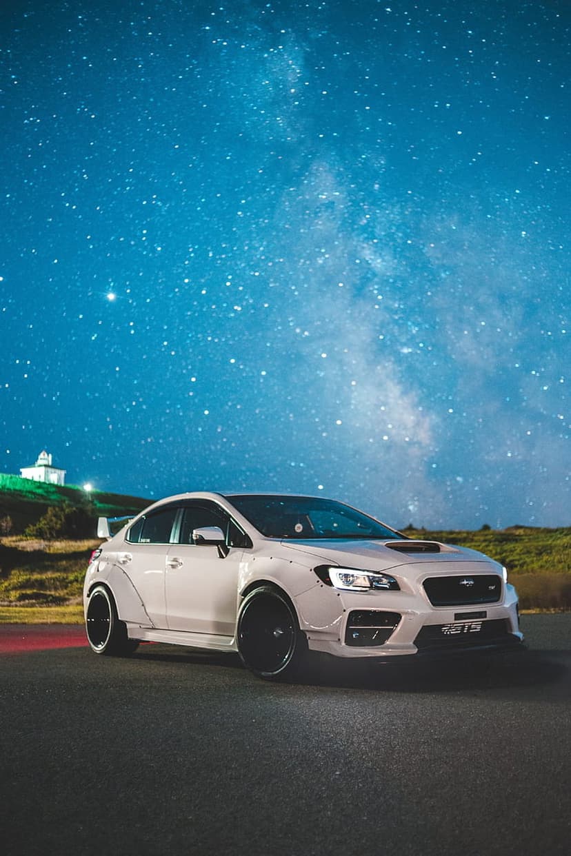 White widebody Subaru WRX parked on a dark road under a vibrant Milky Way galaxy and a star-filled night sky.