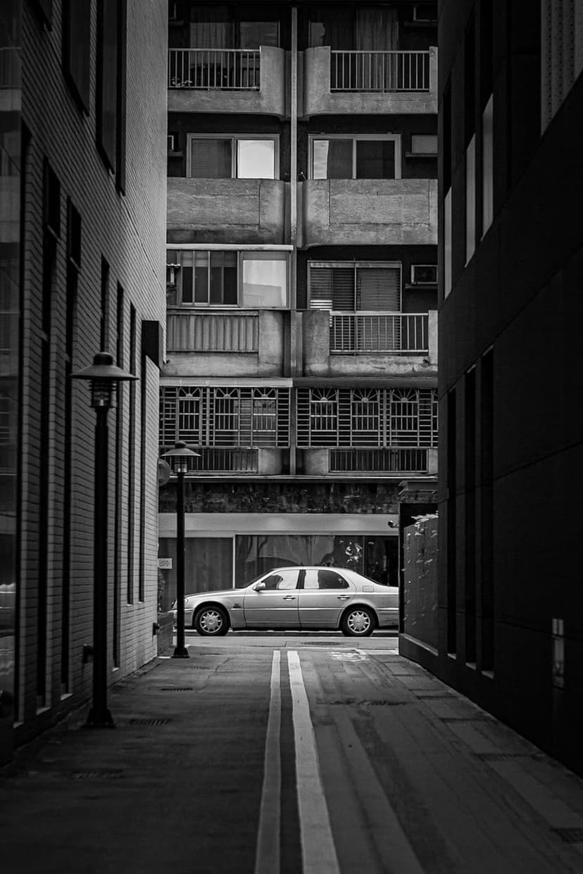 Silver sedan parked in a high-contrast black and white alleyway between tall buildings with textured balconies.