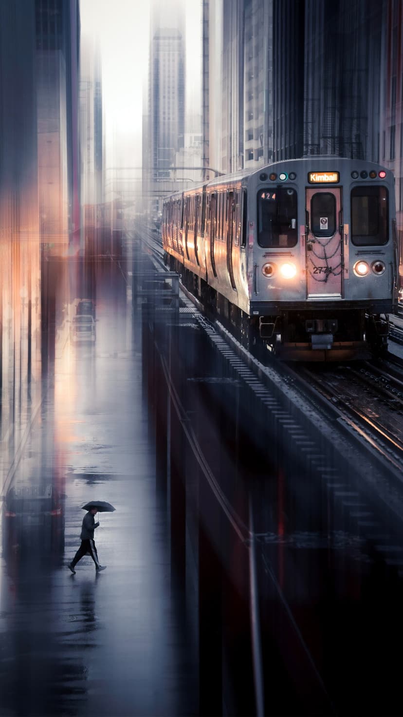 Person with an umbrella crosses a wet street beneath a moving elevated train and towering city skyscrapers.