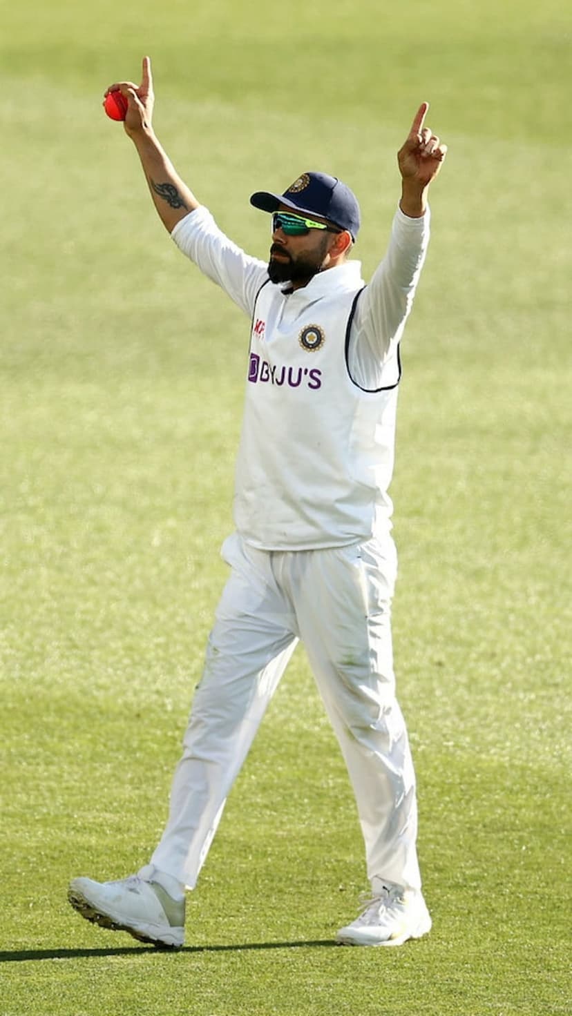 Virat Kohli raises his arms in white cricket gear while holding a red ball against a blurred green field.