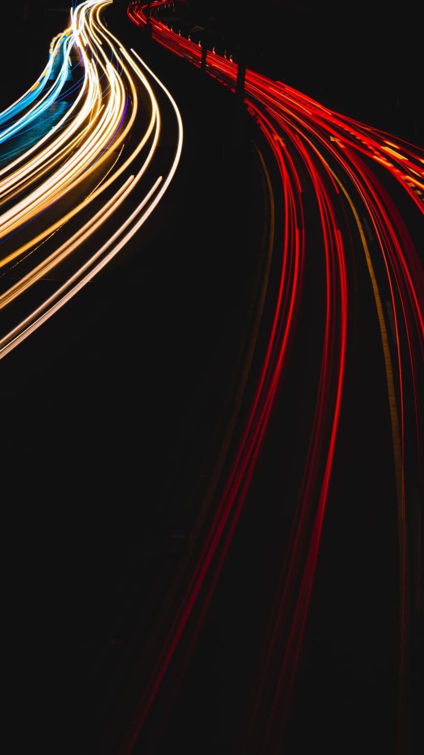 Red and white light trails stretch along a dark asphalt road at night with distant city lights in the background.