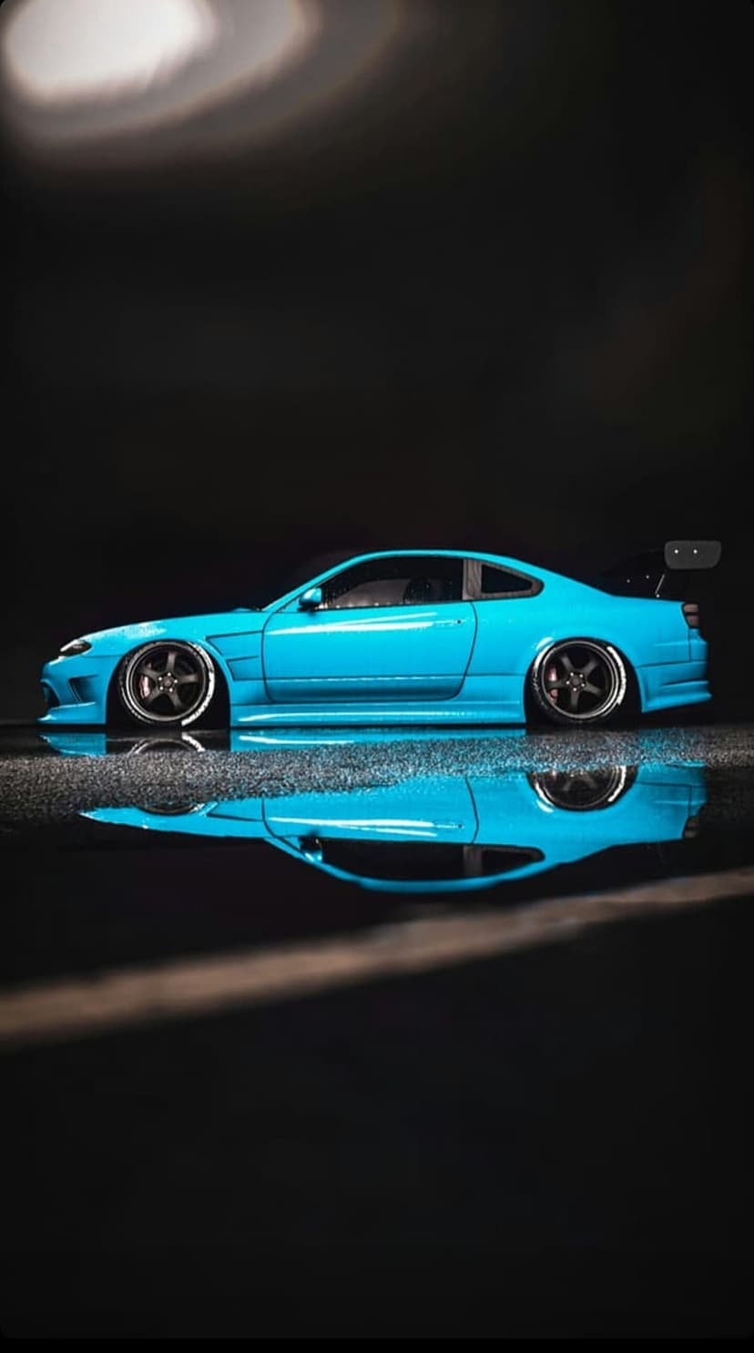 Blue Nissan S15 Silvia sports car parked on a wet pavement with a clear mirror reflection and dark background.
