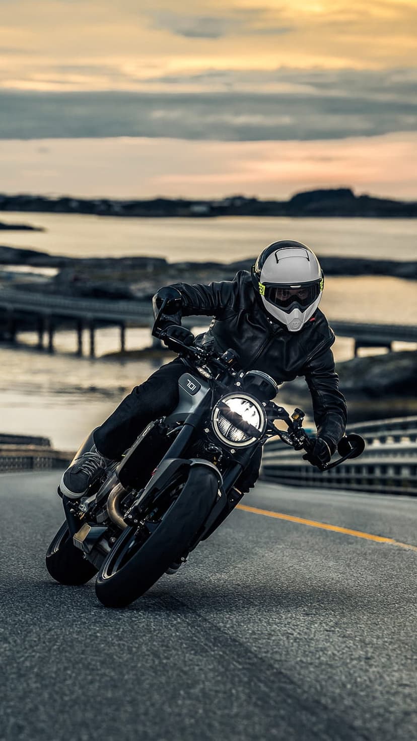 Motorcyclist in a white helmet and black jacket leans into a road curve against a vibrant sunset and bridge.