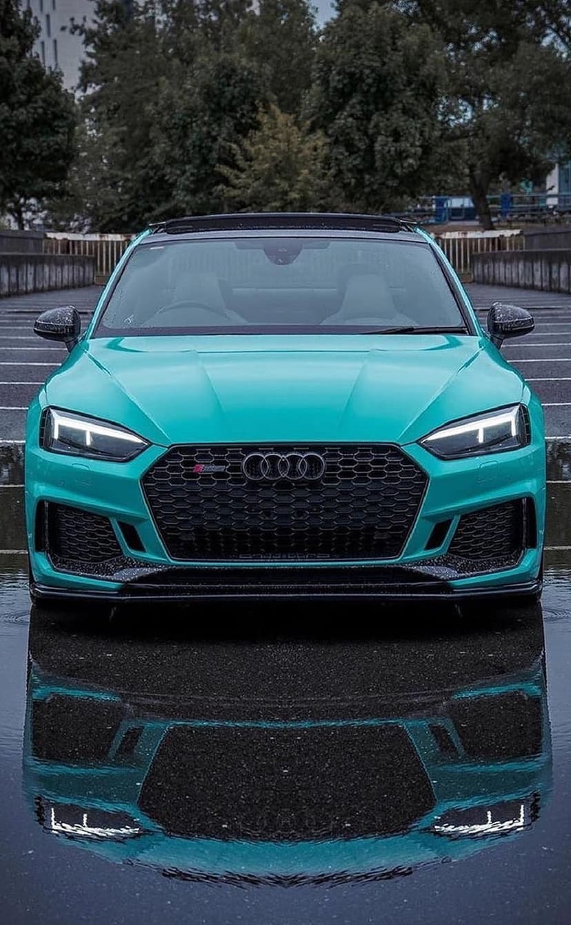 Turquoise Audi RS5 sports car on reflective wet asphalt with an aggressive front grille and glowing LED lights.