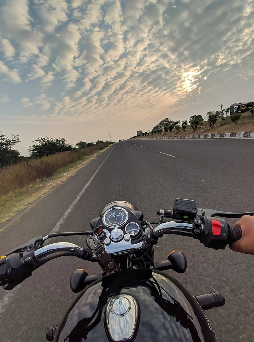 Motorcyclist POV riding a black bike on a desert highway under a dramatic sunset sky with rippling orange clouds.