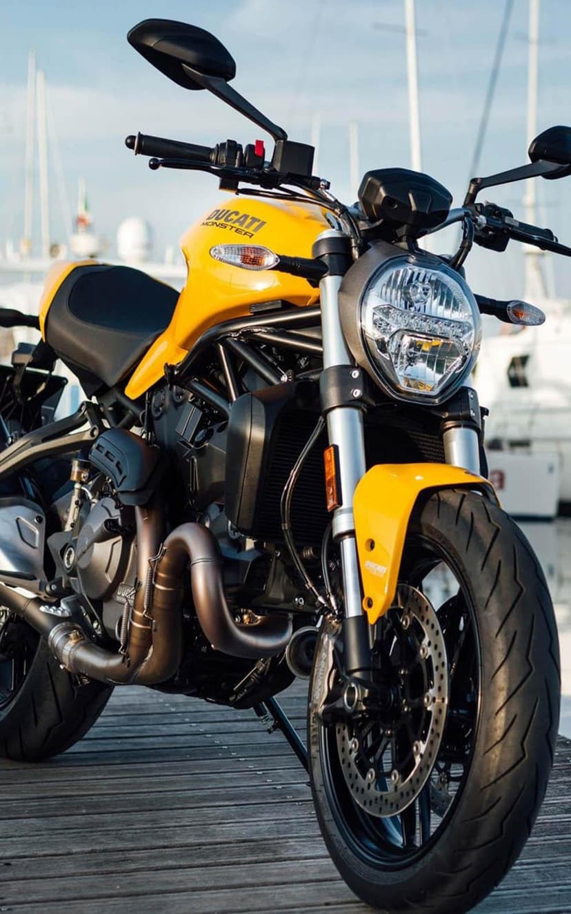 Yellow Ducati Monster motorcycle parked on a wooden pier with blurred sailboats and a blue sky background.