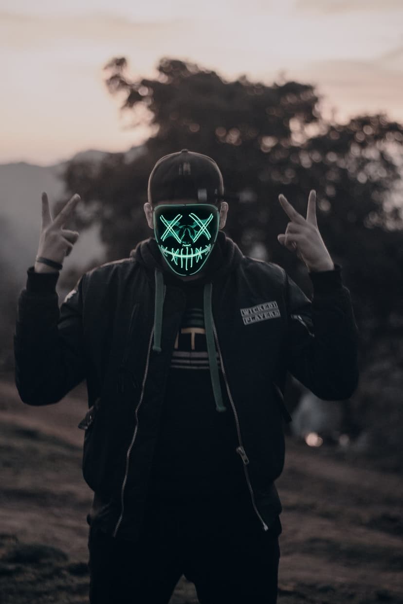 Man in a glowing green LED mask wearing a black jacket making peace signs against a twilight forest backdrop.