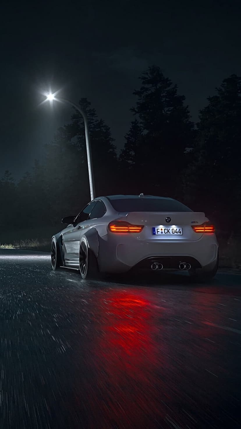 White BMW M4 coupe with widebody kit driving on wet asphalt at night under red taillight reflections.