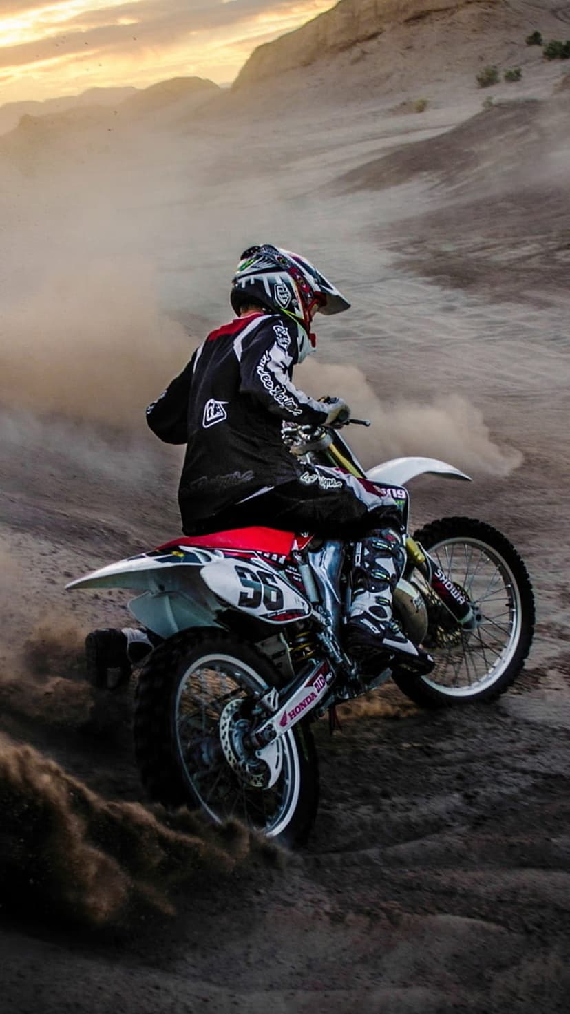 Motocross rider on a red and white dirt bike kicks up a cloud of sand during a sunset race in hilly terrain.