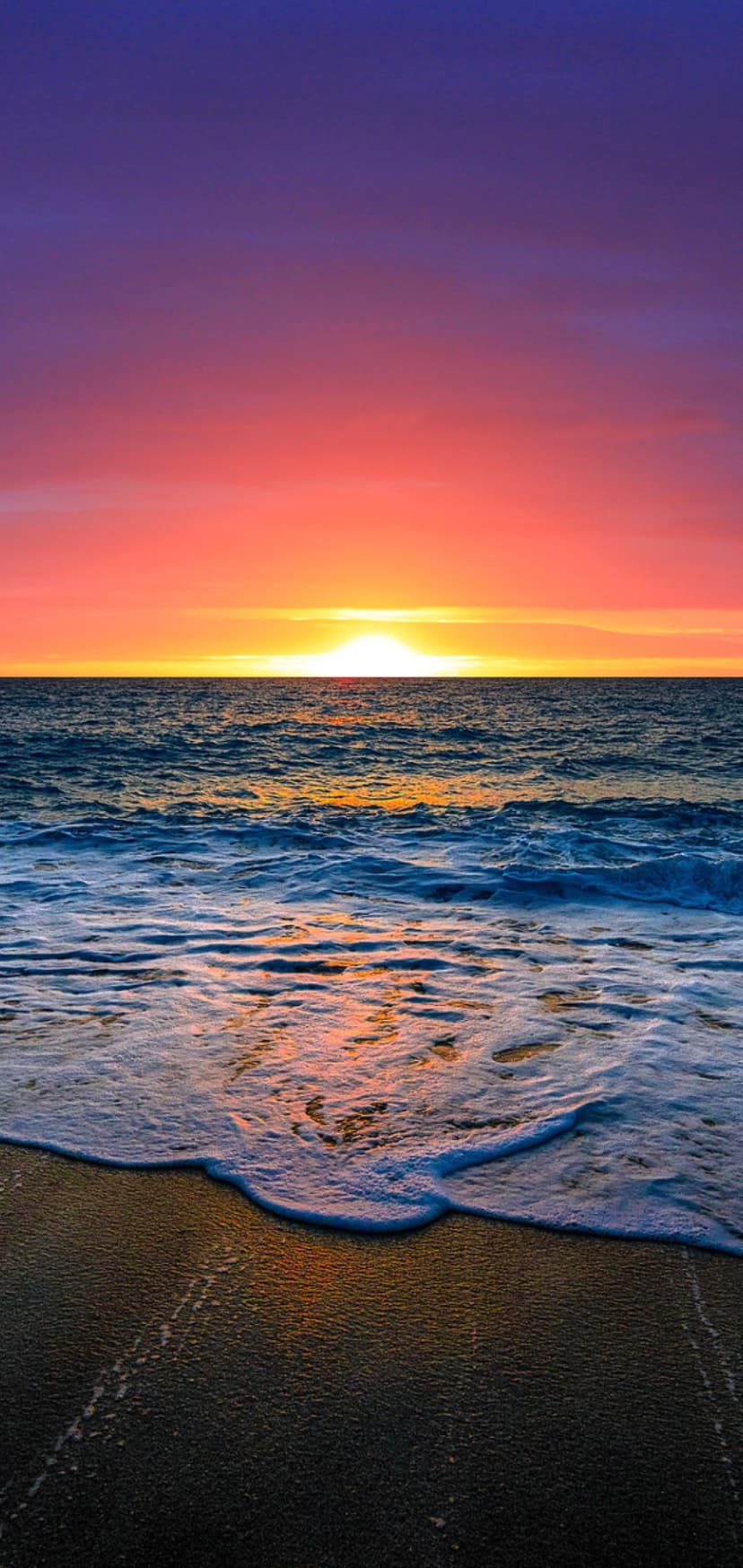 Fiery orange and purple clouds hang over white-capped ocean waves washing onto a dark sandy beach at twilight.