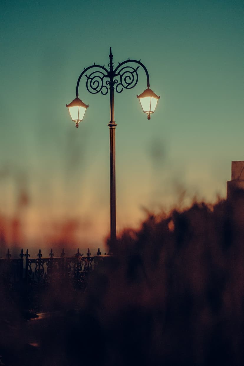 Double-headed ornate street lamp with glowing lanterns against a teal and orange gradient sky during twilight.