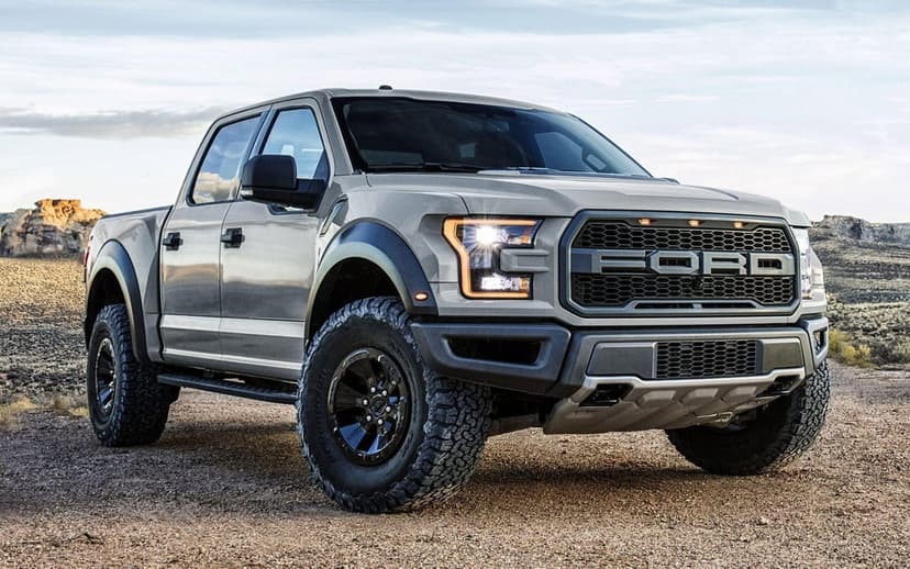 Gray Ford F-150 Raptor pickup truck with off-road tires parked on a rocky desert trail under a bright sky.